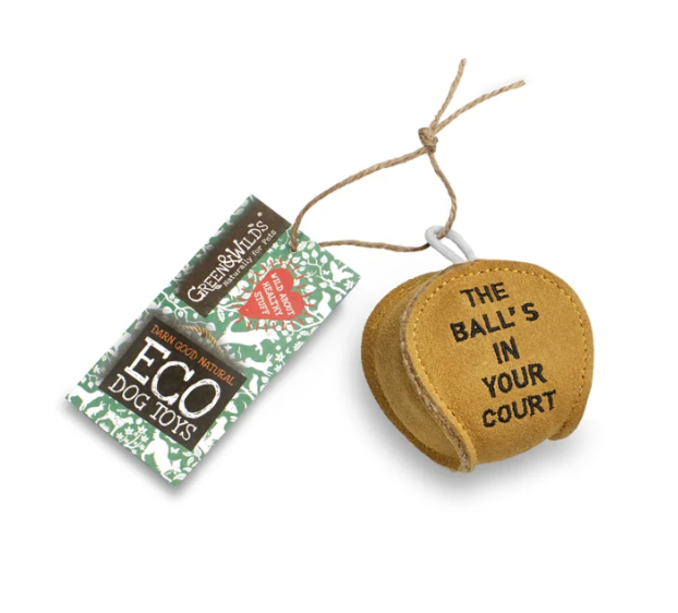 A round tan dog toy with black text reading 'THE BALL'S IN YOUR COURT' on the front, shown next to its branded paper hang tag on a white background.