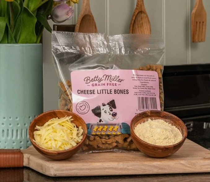 A bag of Betty Miller Grain Free Cheese Little Bones dog treats sits on a wooden cutting board, styled with bowls of shredded cheese and dry ingredients in a kitchen setting.