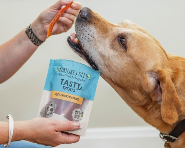 Dog sniffing a package of Natures Deli Tasty Treats soft chicken strips held by a person.