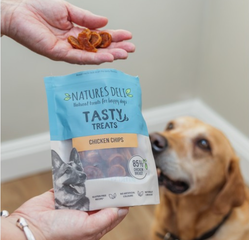 Natures Deli Tasty Treats Chicken Chips package with a hand holding treats and a dog looking on.