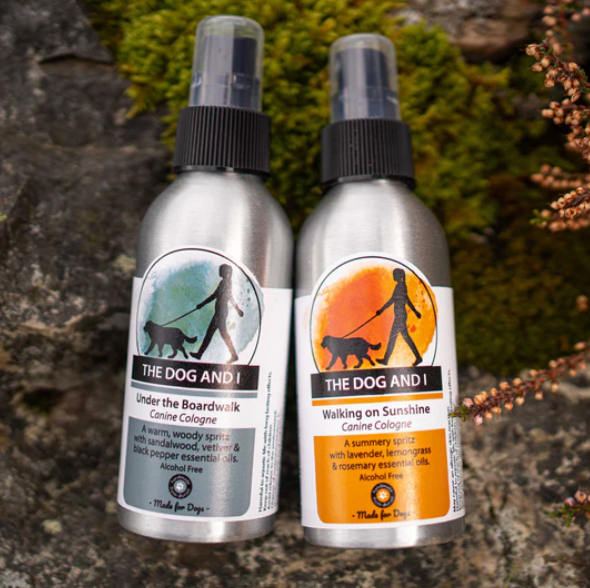 Two spray bottles of dog cologne with labels 'The Dog and I Under the Boardwalk' and 'The Dog and I Walking on Sunshine', set against a natural background with moss and mushrooms.