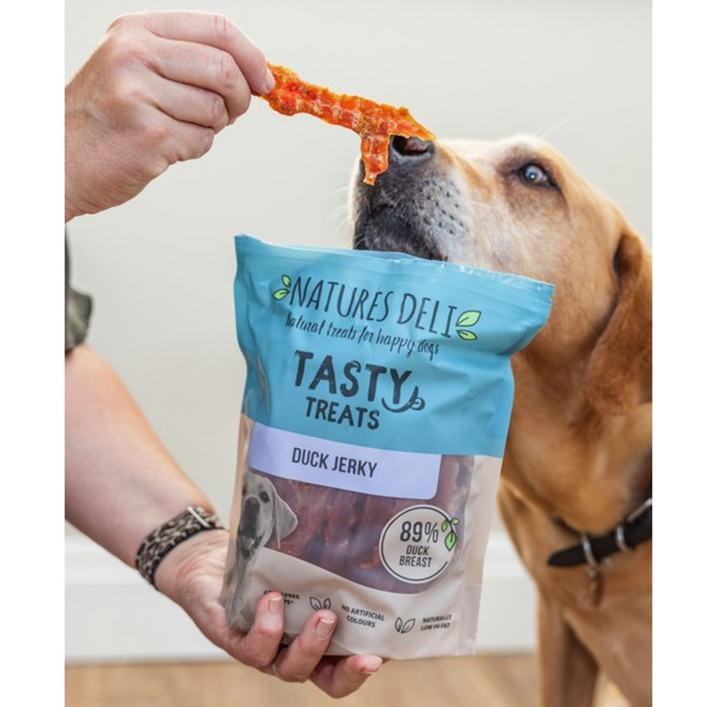Dog eating a treat from a 'Natures Deli Tasty Treats' (duck jerky) package with a visible brand logo.