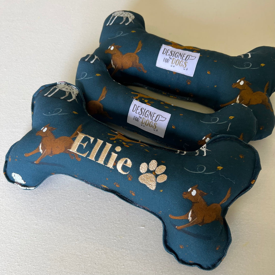 Three dog bone toys, one embroidered with the name Ellie on it, all in the Emily Cole Muddy Paws fabric (dark blue fabric with dogs running, leaving behind muddy paw prints)