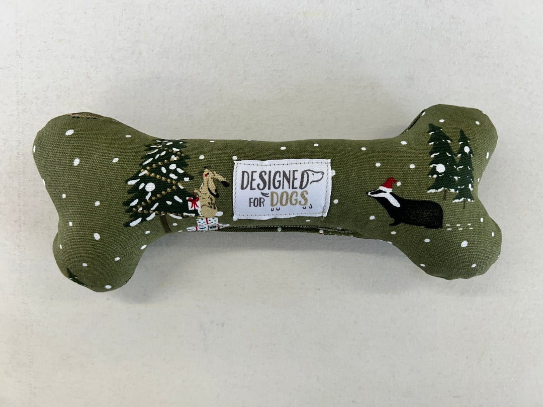 An olive green fabric dog toy shaped like a bone, decorated with a winter scene featuring pine trees, snow, and embroidered dogs. A central white patch reads 'DESIGNED FOR DOGS'.