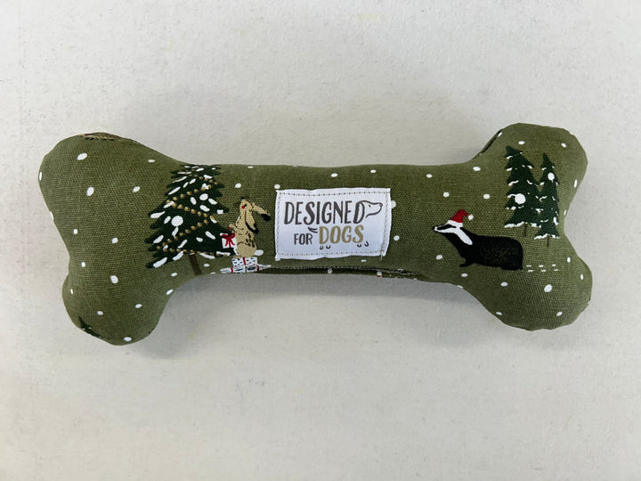 An olive green fabric dog toy shaped like a bone, decorated with a winter scene featuring pine trees, snow, and embroidered dogs. A central white patch reads 'DESIGNED FOR DOGS'.