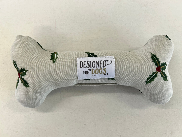 A bone-shaped dog toy made of light beige fabric with green holly leaves and red berries, featuring a central white label that reads 'DESIGNED FOR DOGS.'.