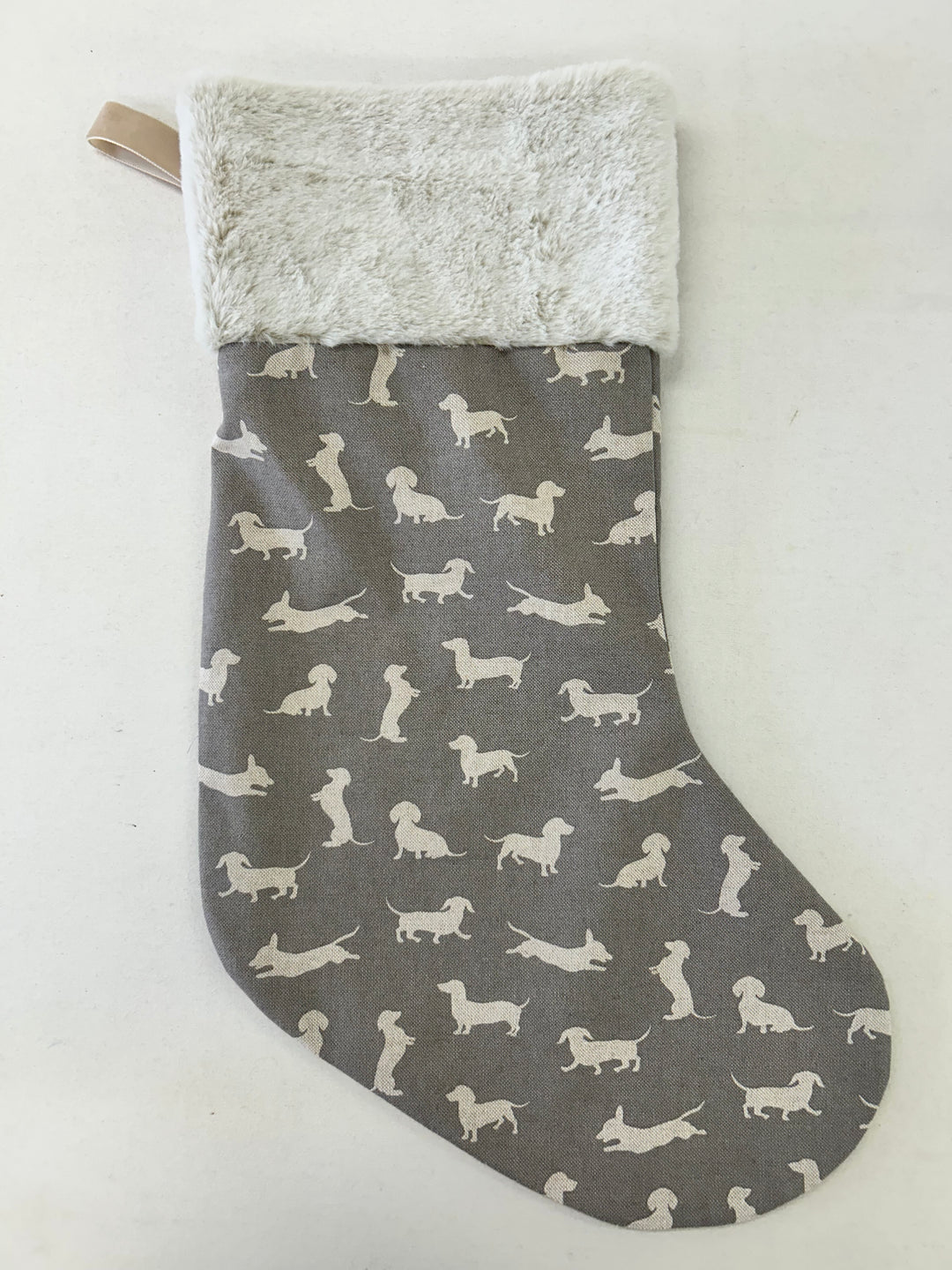 A Christmas stocking with a gray fabric body printed with white dachshund dog silhouettes, featuring a fluffy cream-colored faux fur cuff and a small beige hanging loop.