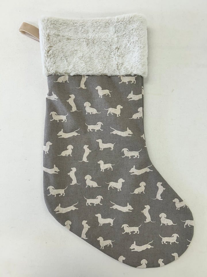 A Christmas stocking with a gray fabric body printed with white dachshund dog silhouettes, featuring a fluffy cream-colored faux fur cuff and a small beige hanging loop.