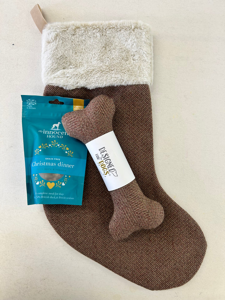A brown herringbone Christmas stocking with a cream faux-fur cuff, displayed with a bone-shaped dog toy and a blue bag of The Innocent Thing Christmas Dinner dog treats on a light beige background.