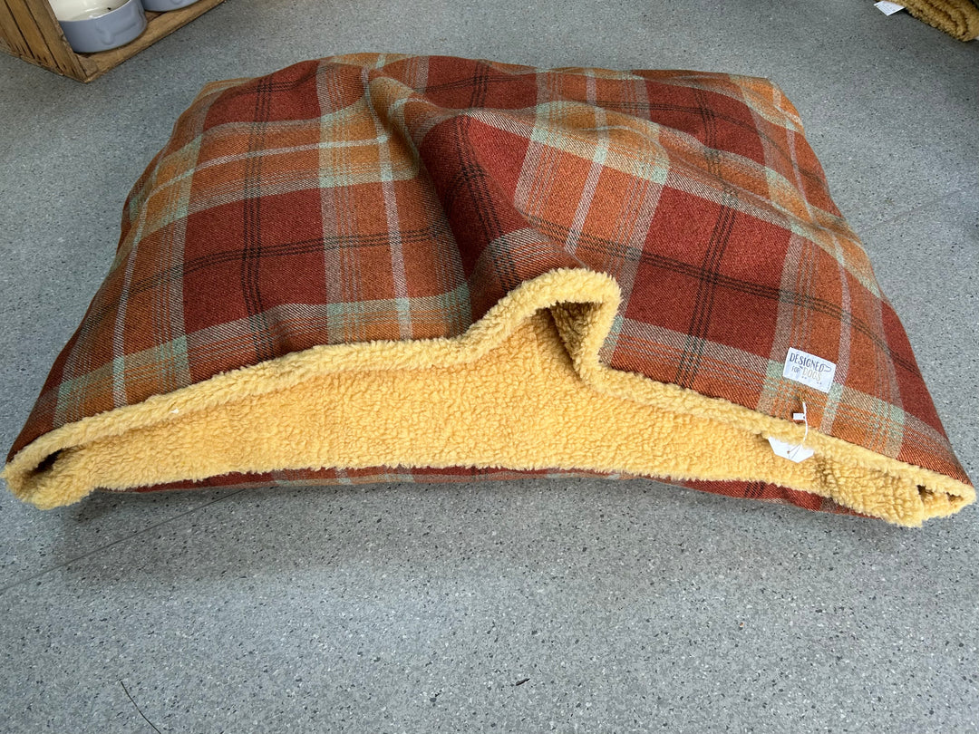 A square pet bed with a rust-orange and beige plaid patterned fabric exterior and a soft yellow sherpa lining, shown on a gray concrete floor.
