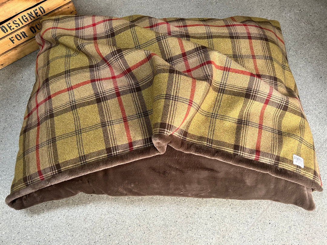 A square pet bed with a yellow, brown, and red plaid fleece cover and a plush brown suede-like underside, shown on a speckled gray floor with a wooden crate partially visible in the top left.