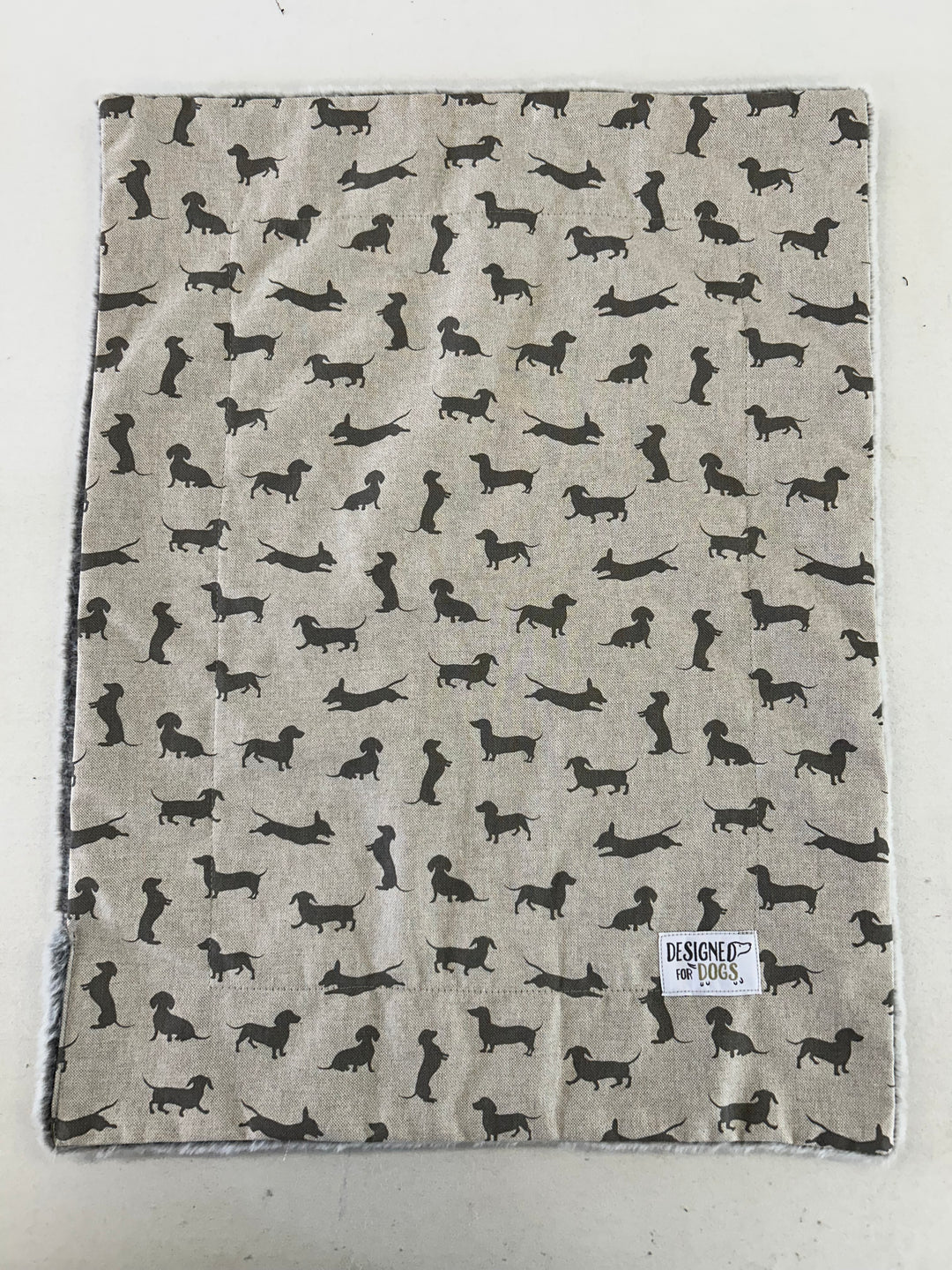 A square fabric item, likely a blanket or towel, featuring a repeating pattern of black dachshund dog silhouettes on a light beige linen-like background. A small white label reading 'HESPOE of HOUS' is visible in the bottom right corner.