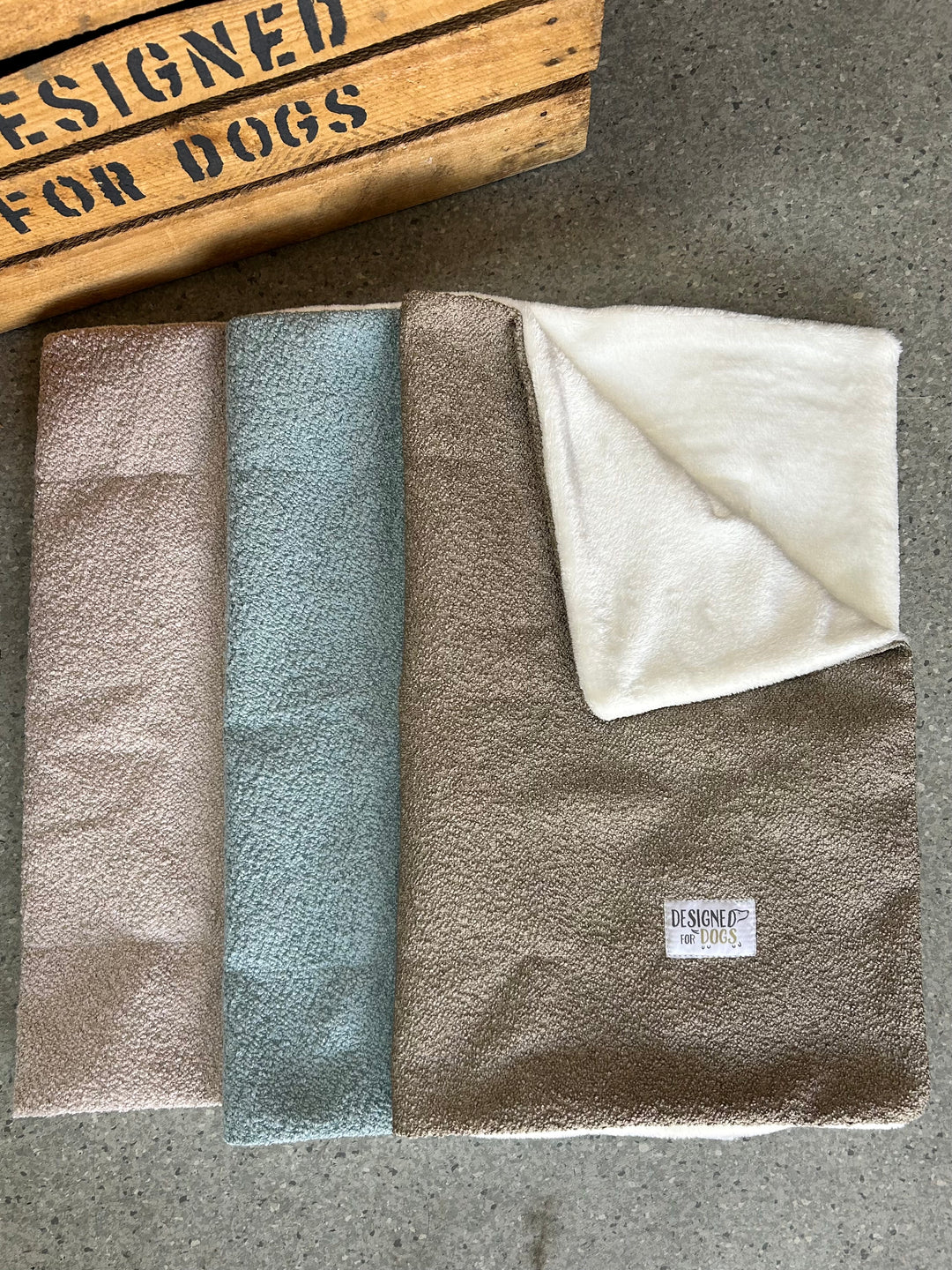 Three folded dog towels in taupe, dusty blue, and cream are arranged on a gray concrete surface next to a wooden crate. The taupe towel is open to show its soft plush reverse side, and a small 'BESPOKE' brand label is visible.