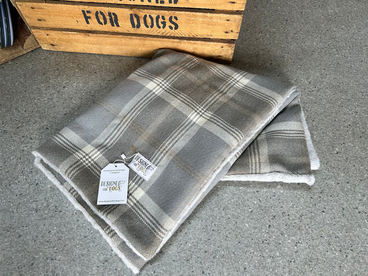 A folded plaid blanket in gray, beige, and white tones with a white tag reading 'OLSONE for dogs', placed on a gray floor in front of a wooden crate.