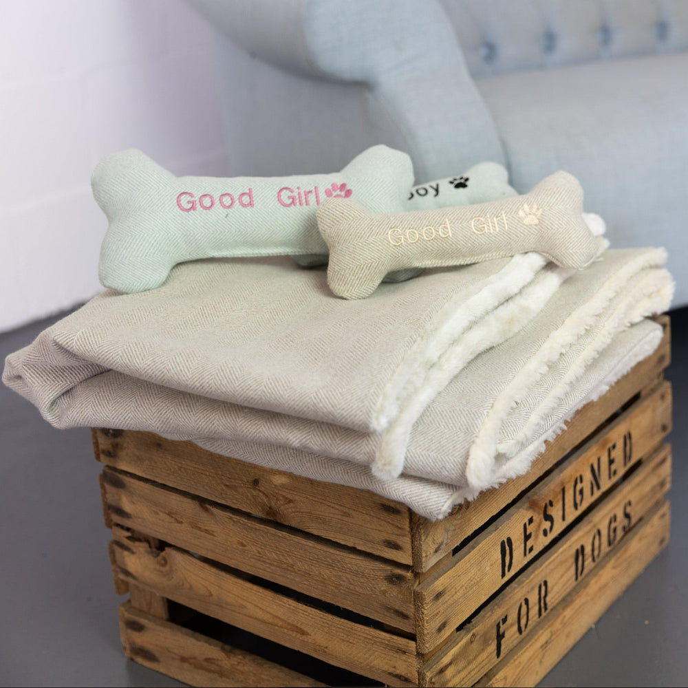 A folded cream-colored blanket or throw is displayed on a rustic wooden crate, with two plush bone-shaped dog toys resting on top. One toy is pale mint green and reads 'Good Girl' with a heart, while the other is cream with 'Good Boy' and paw prints.