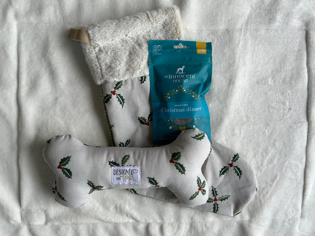 A Christmas-themed pet gift set featuring a white fabric dog stocking with green holly and red berries, a bone-shaped cushion with matching embroidery, and a blue bag of Britoche dog treats labeled 'Christmas dinner' on a white textured blanket.