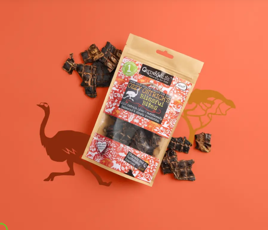 A package of Green & Wild's Ostrich & Emu Biscuit Chews is displayed on a bright orange background with scattered dark brown dog treats around it. The packaging features a gold foil top and bottom with a red and white patterned center, and the brand name Green & Wild's is clearly visible.