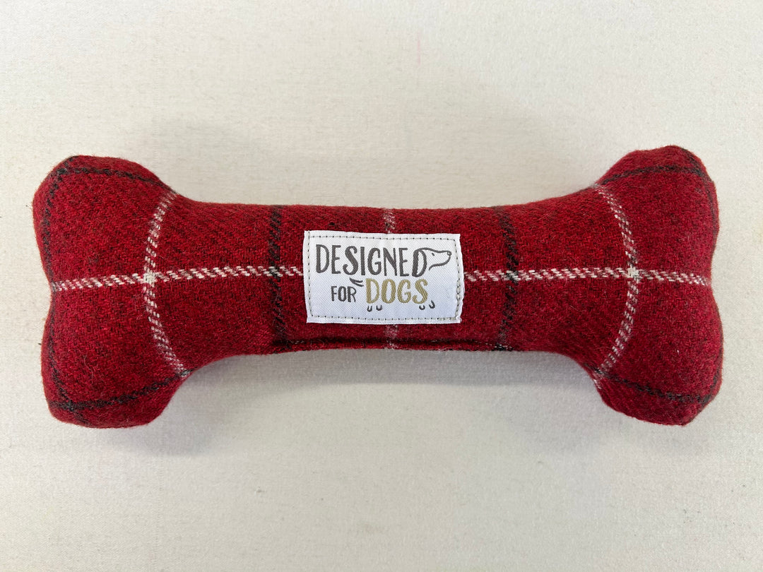 A red plaid dog bone-shaped plush toy with a white label in the center that reads 'DESIGNED FOR DOGS' on a light beige background.
