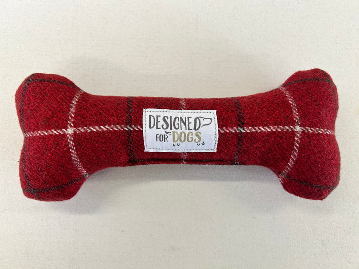 A red plaid dog bone-shaped plush toy with a white label in the center that reads 'DESIGNED FOR DOGS' on a light beige background.