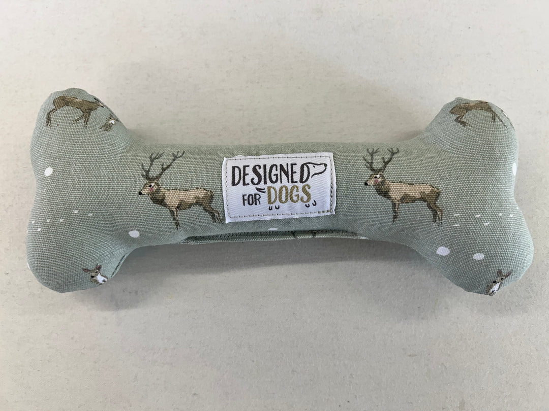 A bone-shaped dog toy made of sage green fabric with embroidered deer and a central white label that reads 'DESIGNED FOR DOGS'.