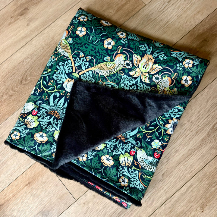 Blanket in the William Morris Strawberry thief blanket on a wooden floor. Fabric shows birds and flowers, with green/blue colours. 