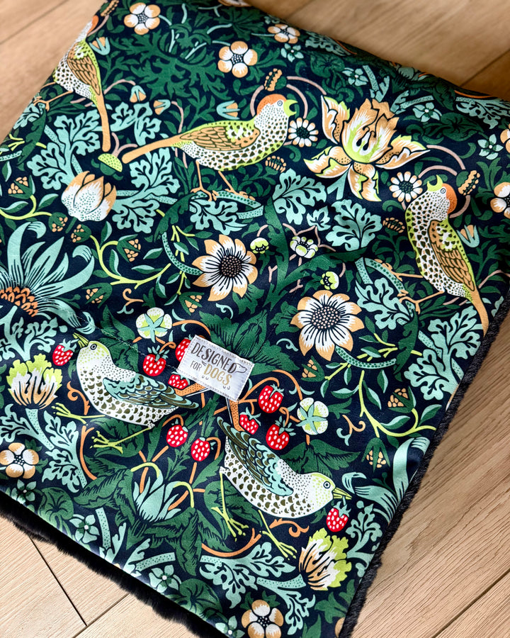 Blanket in the William Morris Strawberry thief blanket on a wooden floor. Fabric shows birds and flowers, with green/blue colours. 