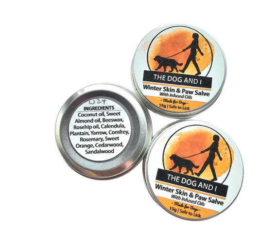 Two travel-sized containers of 'The Dog and I' Winter Skin & Paw Salve with labels indicating almond oil, beeswax, coconut oil, orange, cedarwood, and sandalwood. One container is open showing the product, and the other is closed.
