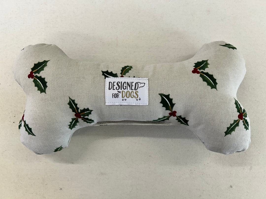 A bone-shaped dog toy made of light beige fabric with a green holly and red berry print, featuring a central white label that reads 'DESIGNED FOR DOGS'.