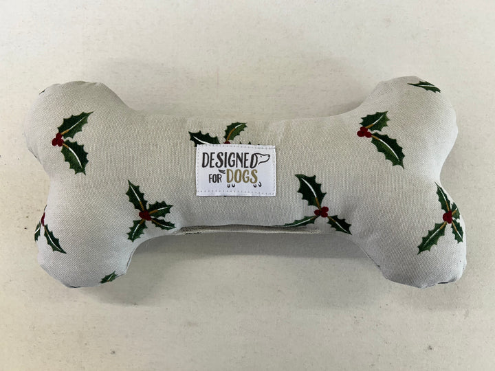 A bone-shaped dog toy made of light beige fabric with a green holly and red berry print, featuring a central white label that reads 'DESIGNED FOR DOGS'.