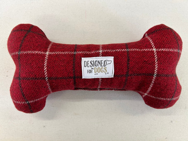 A red tartan dog bone-shaped plush toy with a white label in the center that reads 'DESIGNED FOR DOGS'.