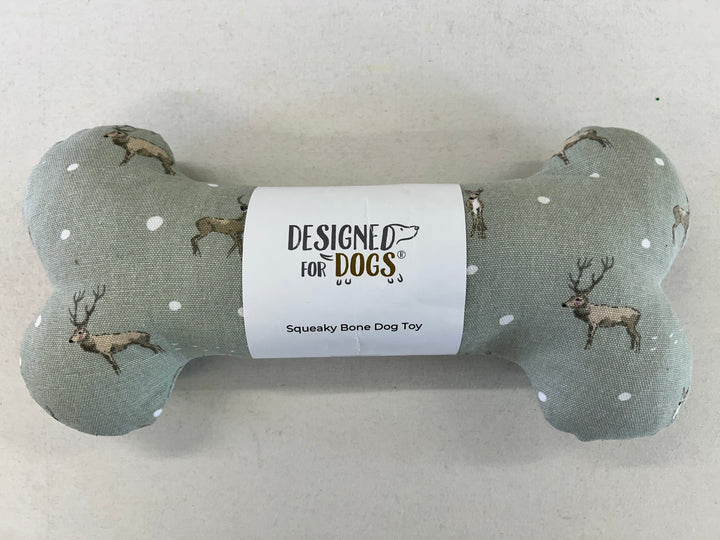 A bone-shaped dog toy made of sage green fabric with a deer and white dot pattern, wrapped in a white paper label that reads 'DESIGNED FOR DOGS' and 'Squeaky Bone Dog Toy'.