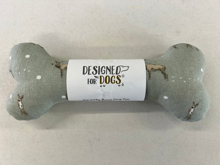 A bone-shaped dog toy made of textured gray fabric with small white dots and printed dog illustrations, wrapped in a white paper band that reads 'DESIGNED FOR DOGS'.