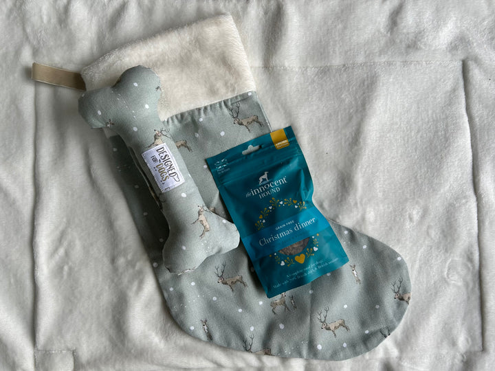 A Christmas-themed dog toy gift set featuring a sage green plush bone toy with white polka dots and a cream fleece cuff, alongside a blue bag of Shimmerlight Labs Christmas treats, laid flat on a white textured blanket.